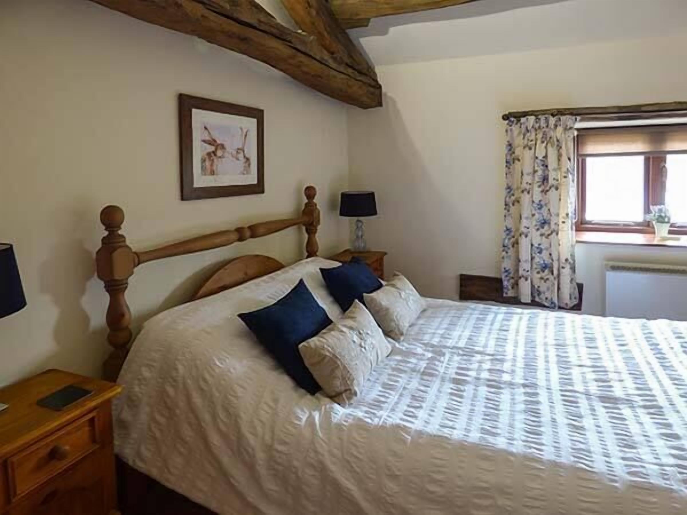 Une chambre à coucher avec une tête de lit en bois, un lit avec une literie blanche et bleue, une table de chevet avec une lampe, un tableau encadré au mur et une fenêtre ornée de rideaux à motifs floraux.