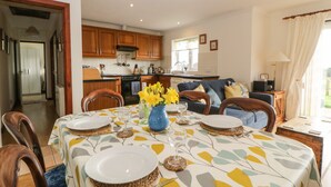 A dining area with a table set for two, a kitchen in the background, and a living area with a sofa and a lamp. - Broom Cottage (Sidmouth)