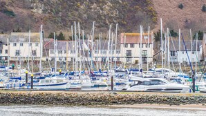 A marina with multiple boats docked, surrounded by buildings and a hilly landscape. - The Moorings (Conwy)