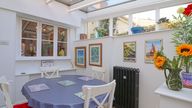 Ein Essbereich mit einem runden Tisch, der mit einer blauen Tischdecke bedeckt ist, weißen Stühlen und einem roten Kissen. An der Wand hängen gerahmte Bilder, ein Heizkörper und ein Fenster mit Blick nach draußen.