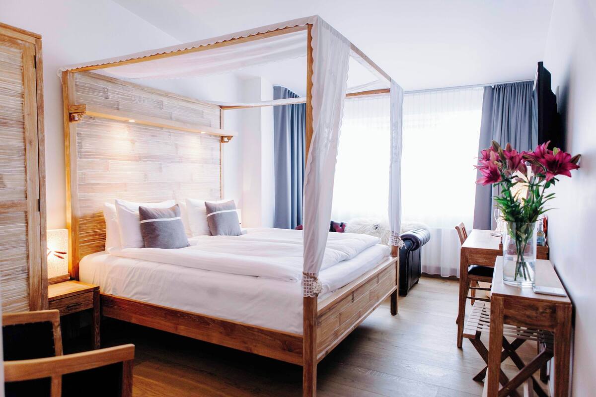 Una cama con dosel de madera y ropa de cama blanca, un escritorio de madera con una silla, y un florero con flores sobre el escritorio.