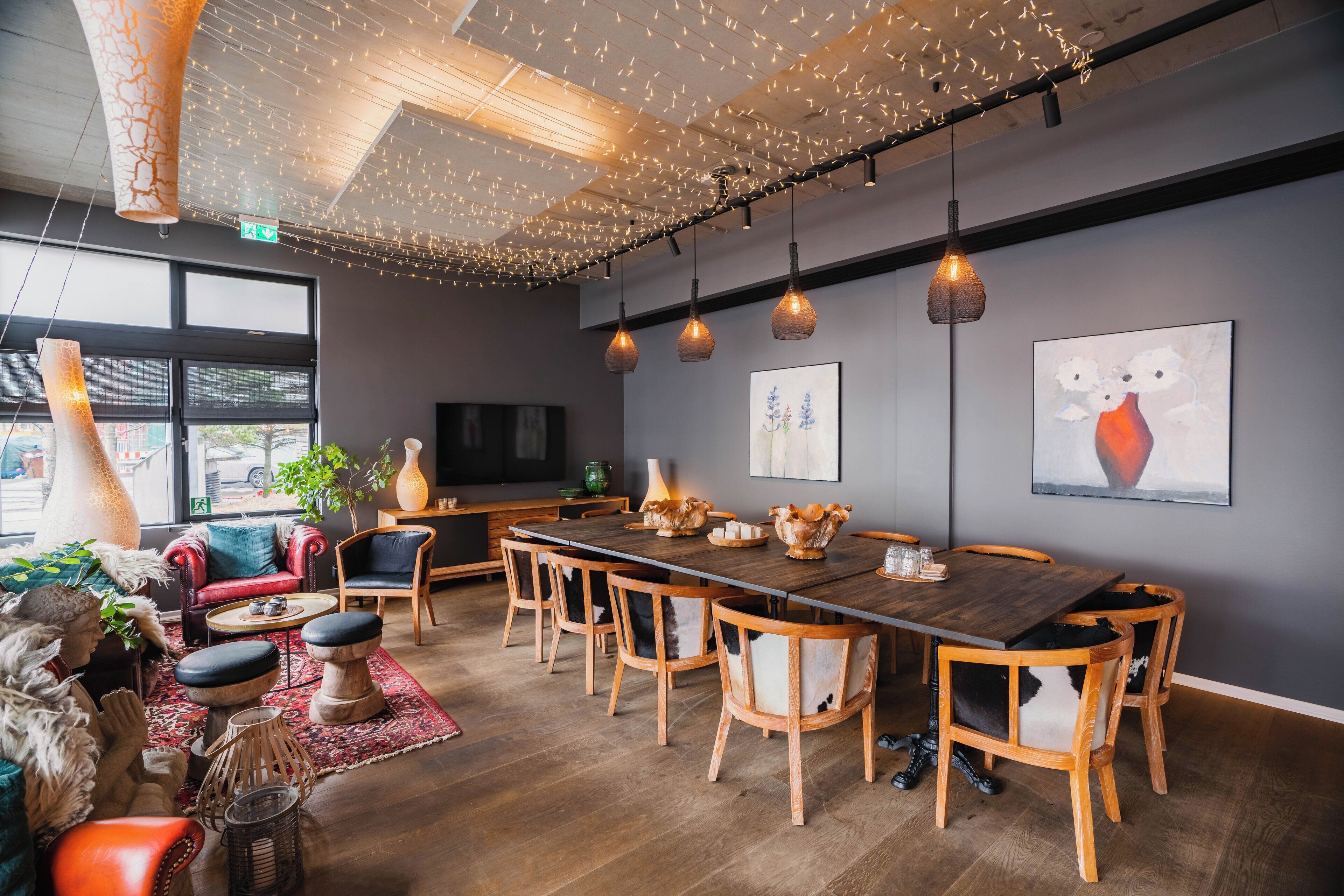 A modern dining area with a long wooden table, multiple chairs, hanging pendant lights, and artwork on the walls.