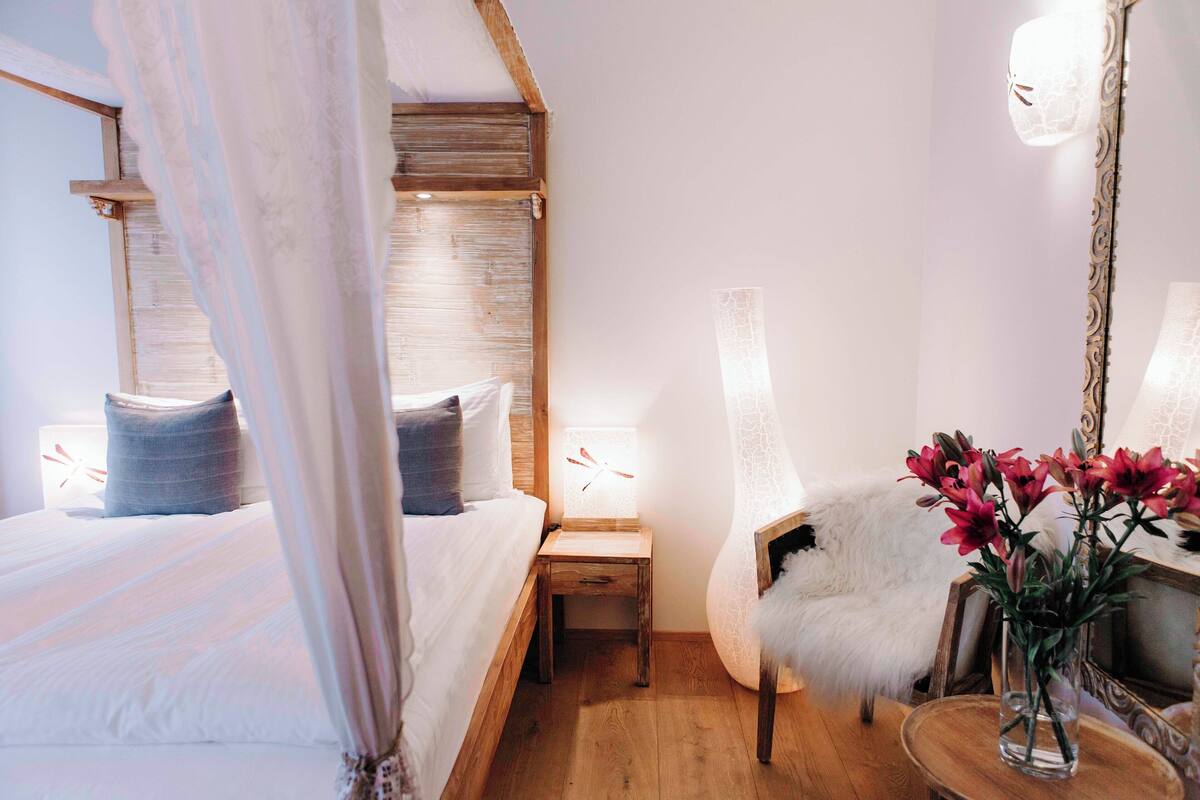 Un dormitorio con cama con dosel, una mesita de noche con lámpara, una silla con una manta de piel sintética y un jarrón con flores.