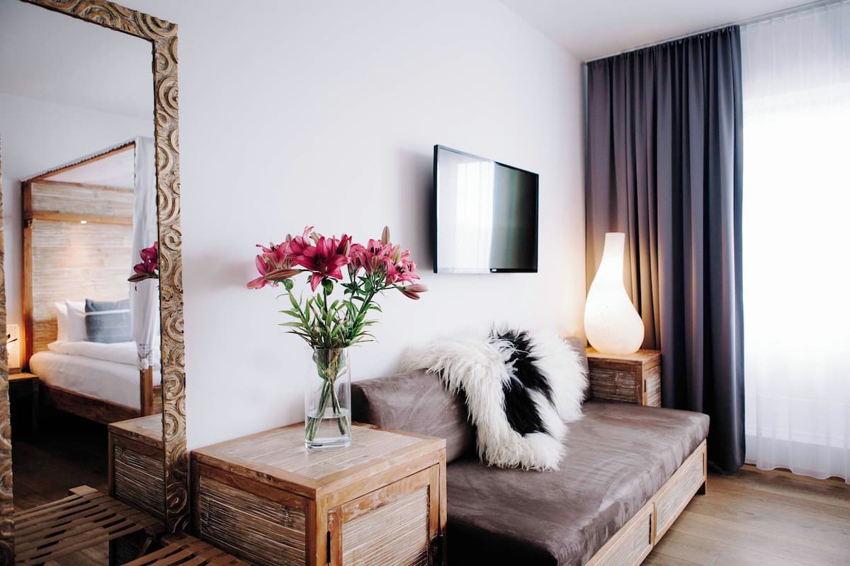 Una habitación de hotel moderna con una mesita de noche de madera, un espejo grande, un televisor de pantalla plana, un sofá con un cojín mullido y un jarrón con flores rosas.