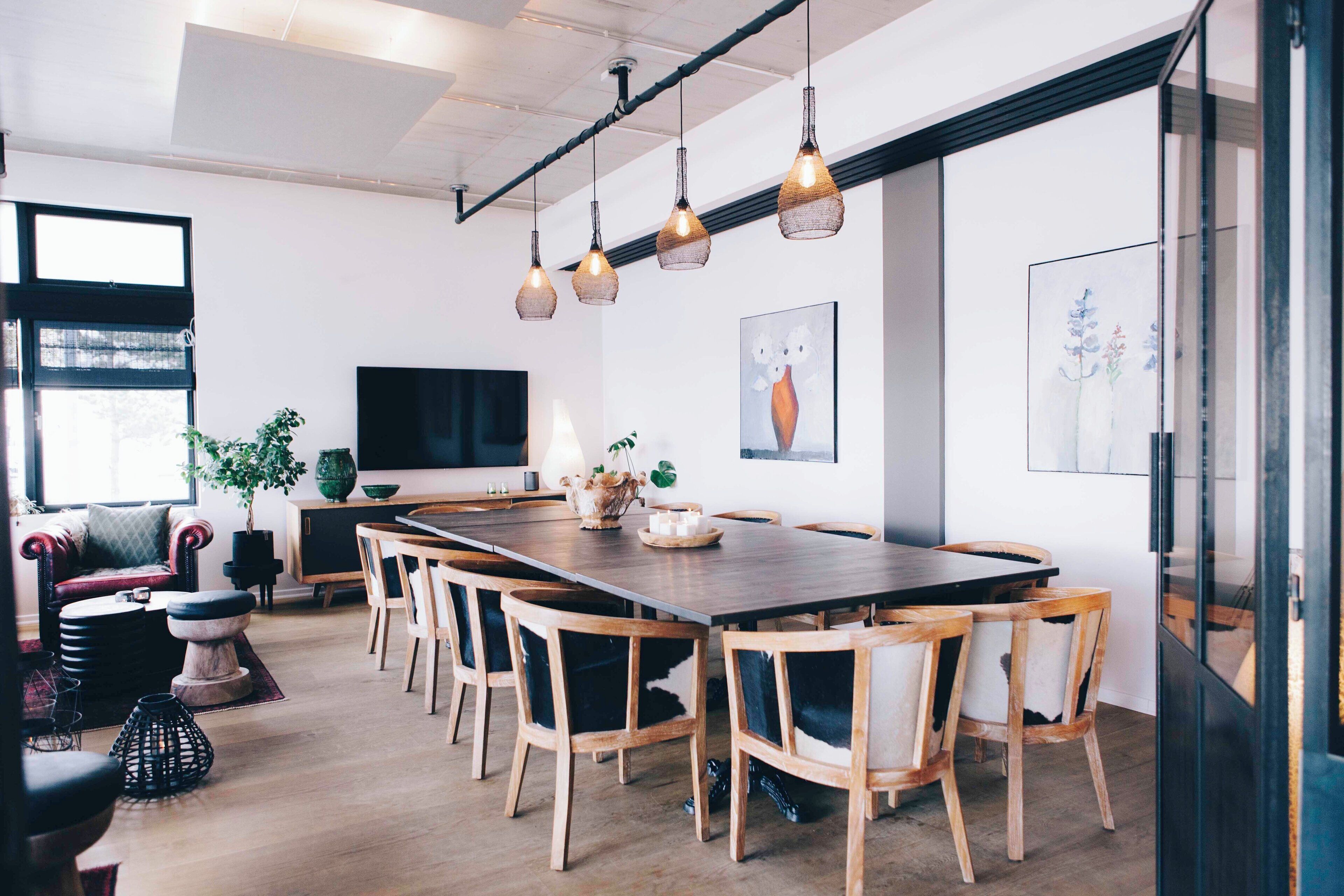Un espace repas moderne avec une longue table en bois, six chaises, des lampes suspendues, un téléviseur et des œuvres d’art accrochées aux murs.