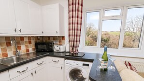 A compact kitchen with white cabinets, a washing machine, a toaster, and a window with a view of trees. - Stable Cottage (Newport)