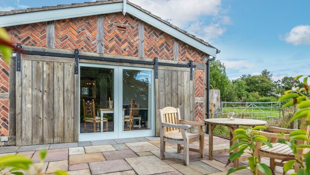 A rustic building with a herringbone brick exterior, large wooden doors, and a stone patio with outdoor seating. - The Lodge (Chester)