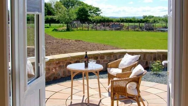 A patio with a table and two wicker chairs, a bottle of wine, and a view of a vineyard. - Byre Cottage (Shrewsbury)