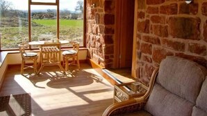 A cozy room with a stone wall, a wooden table with chairs, and a view of a green landscape through large windows. - Byre Cottage (Shrewsbury)
