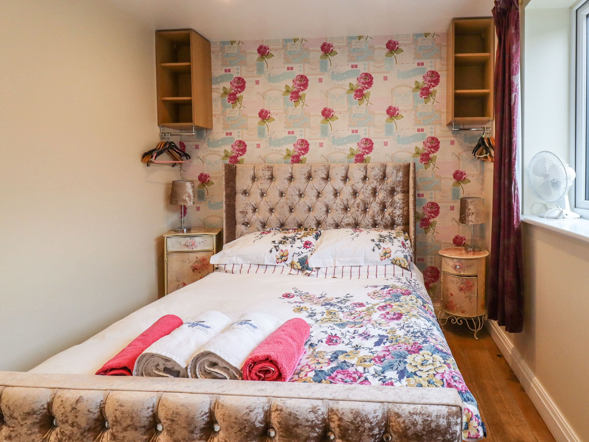 A bedroom with a floral bedspread, a tufted headboard, a wooden wardrobe, and a bedside table with a lamp.