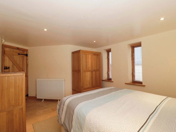 Une chambre à coucher comprenant un lit, une armoire en bois, un radiateur et deux fenêtres équipées de stores.