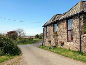 Traditional farmhouse with a single-story design, visible entrance, and window with a curtain, set amidst lush greenery and a fence.