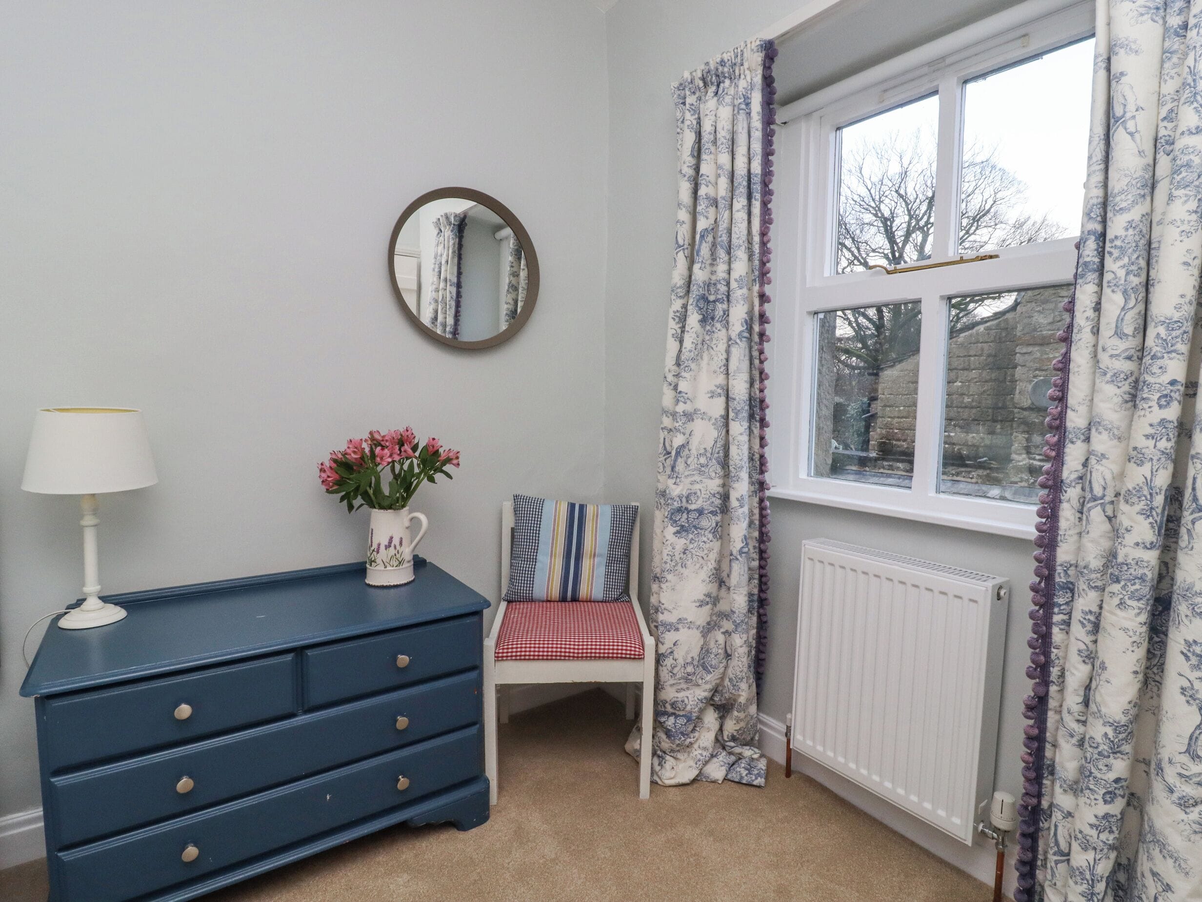 A bedroom with a blue dresser, a white radiator, a window with floral curtains, a mirror, and a chair with a checkered cushion.