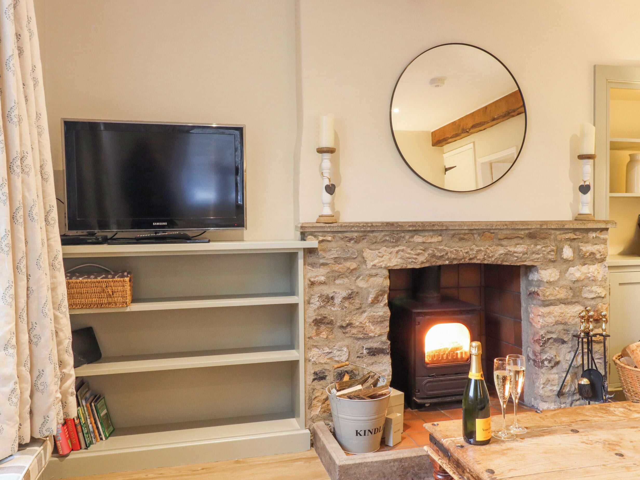 A living room with a television on a shelf, a stone fireplace with a log fire, a mirror above the fireplace, and a table with a bottle and glasses.