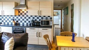 A compact kitchen with a microwave, stove, and checkered backsplash. There is a dining area with a table and chairs, and a sofa with a cushion. - Crud yr Awel (Y Felinheli)