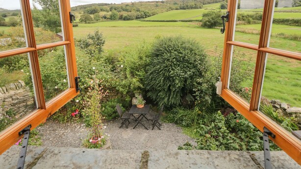 Une vue depuis une fenêtre à cadre en bois, donnant sur un paysage verdoyant avec un mur de pierre, une petite table et des chaises.