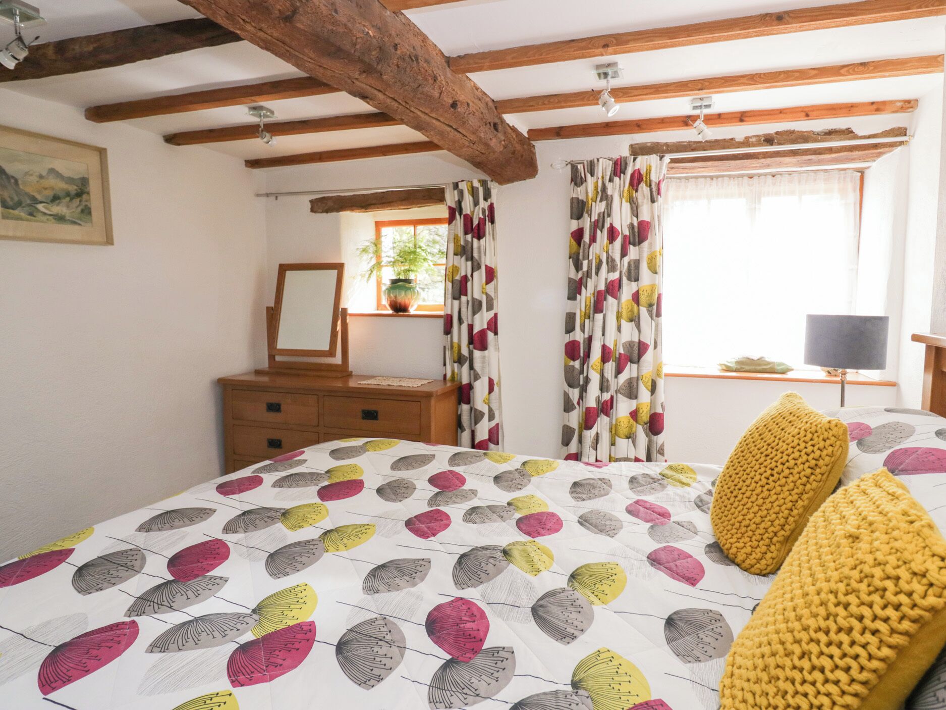 Un dormitorio con una cama con colcha de estampado floral, vigas de madera en el techo, un aparador, un espejo y una ventana con cortinas.