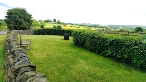 A stone wall, a wooden bench, and a black trash bin in a grassy area with a stone wall and a hedge.