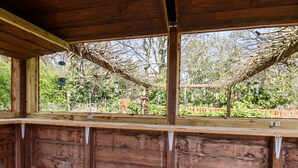 Eine Holzhütte mit einer Bank und einem Tisch, ein Fenster mit Blick auf Bäume und einem Zaun, sowie hängende Laternen.