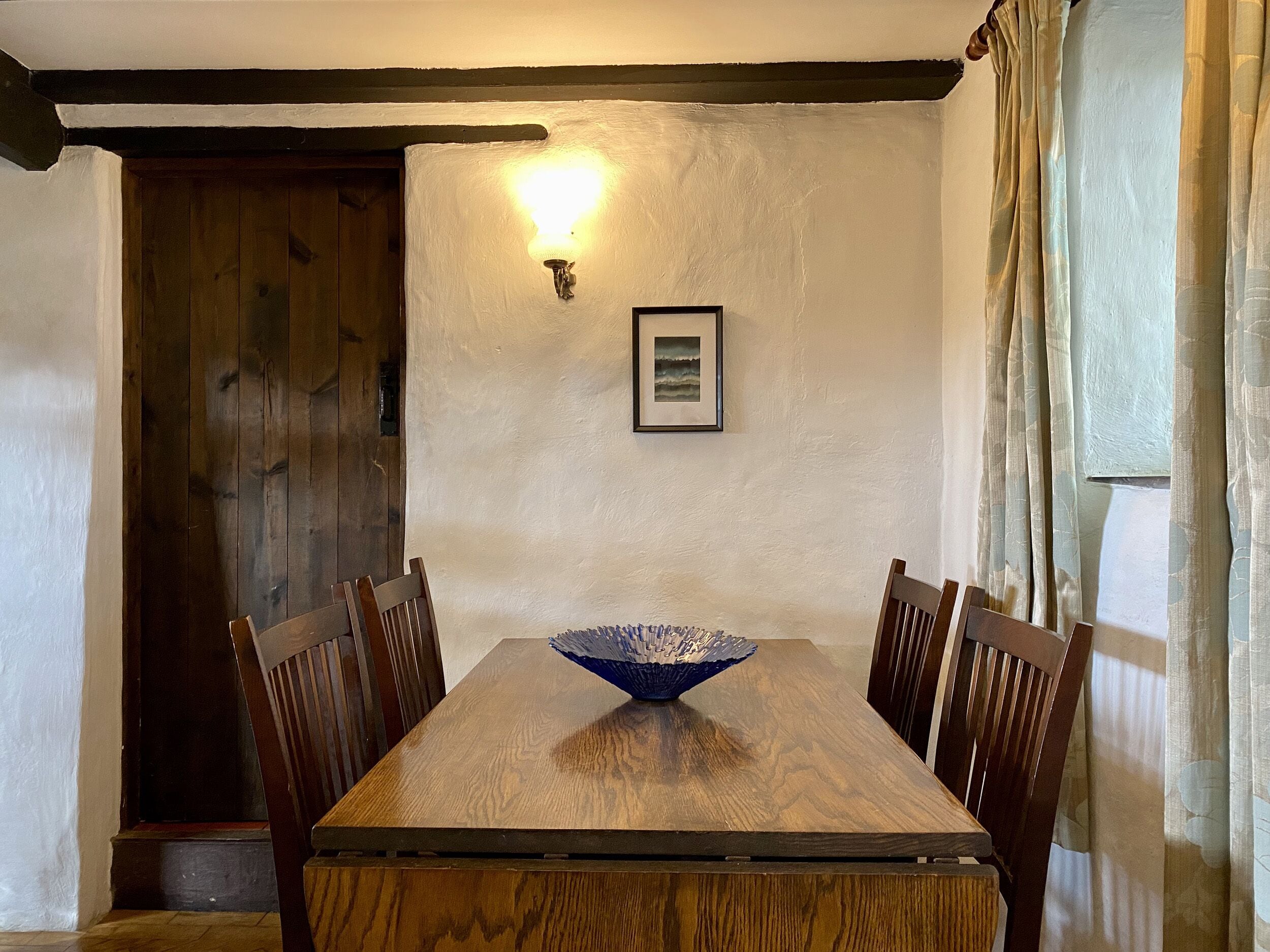 Ein Esszimmer mit Holztisch, Stühlen, einer Wandleuchte, einem gerahmten Bild und einer Tür zu einem Holzinneren.