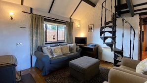 A living room with a sofa, ottoman, and a television. There is a window with curtains, a staircase, and a wall-mounted light fixture. - Hendy Cottage (Pwllheli)