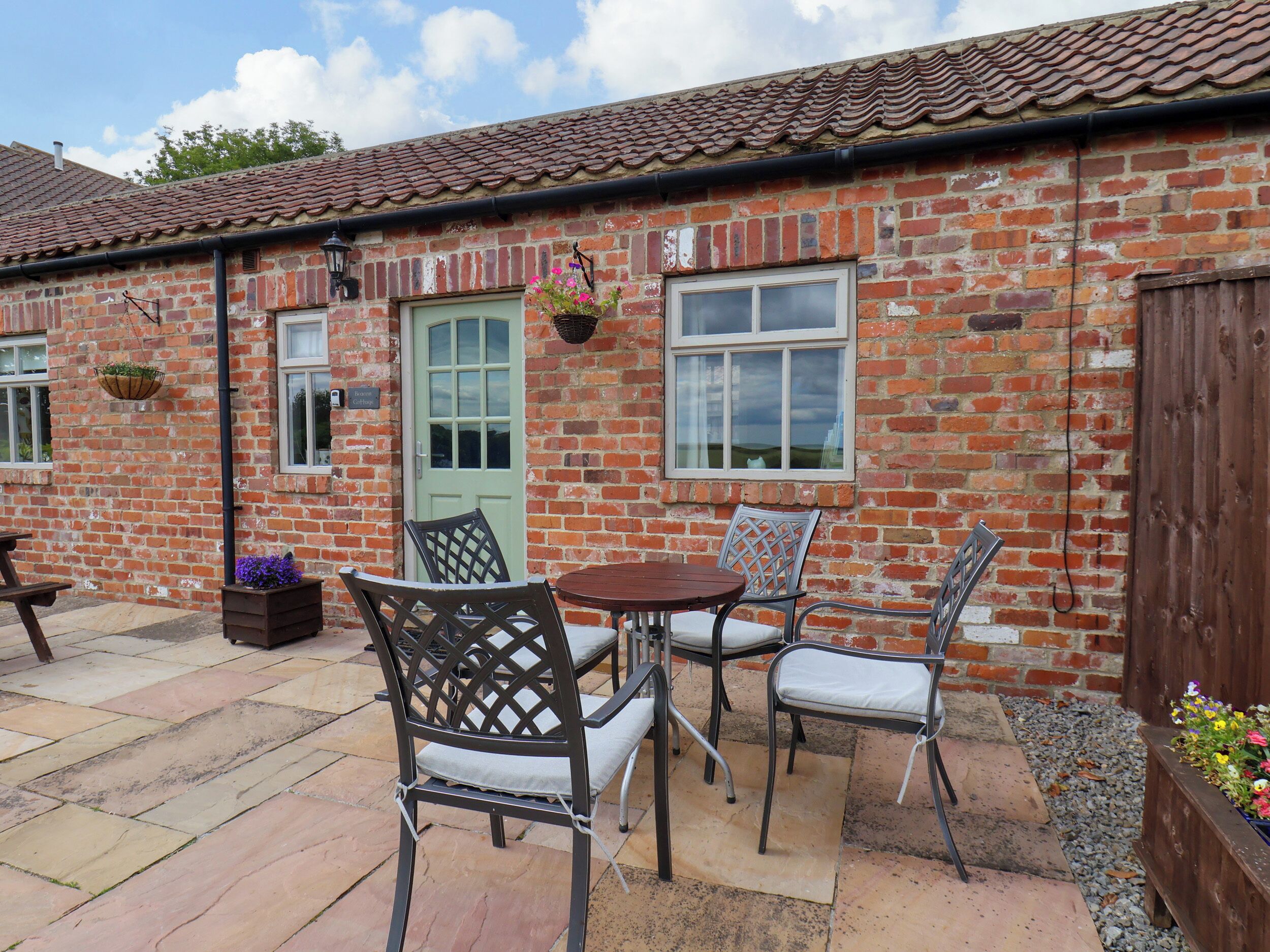 A brick building with a tiled roof, outdoor seating with metal chairs and a table, and hanging flower baskets.