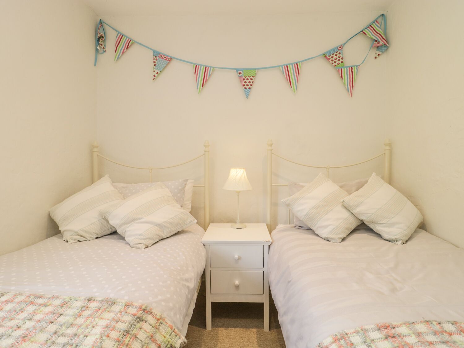 Quarto duplo com duas camas, cada uma com travesseiro e cobertor. Um criado-mudo com abajur fica entre as camas. Uma fileira de bandeirinhas triangulares coloridas pendura sobre as camas.