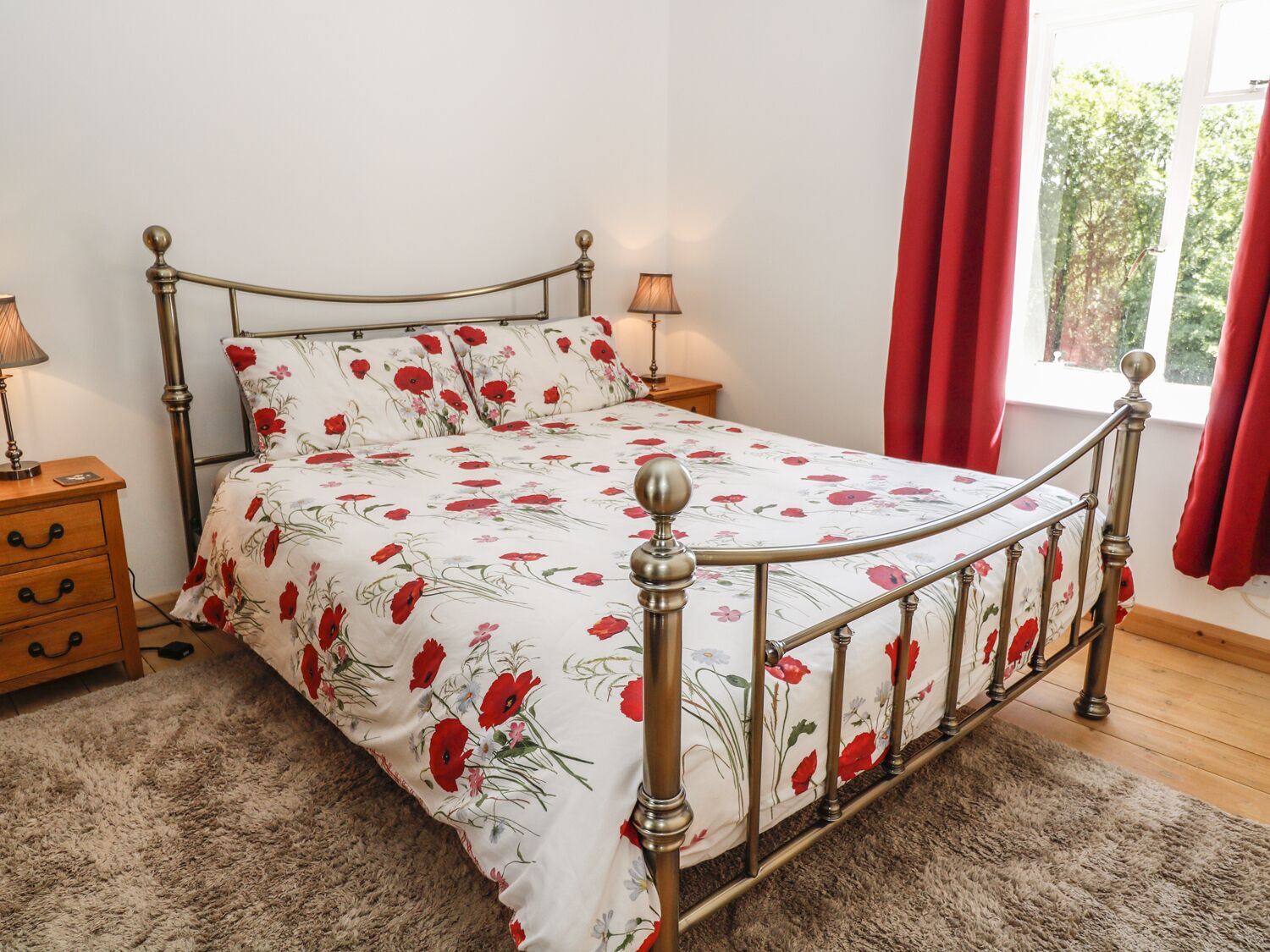 A bedroom with a metal-framed bed, floral bedding, a wooden nightstand, and a window with red curtains.