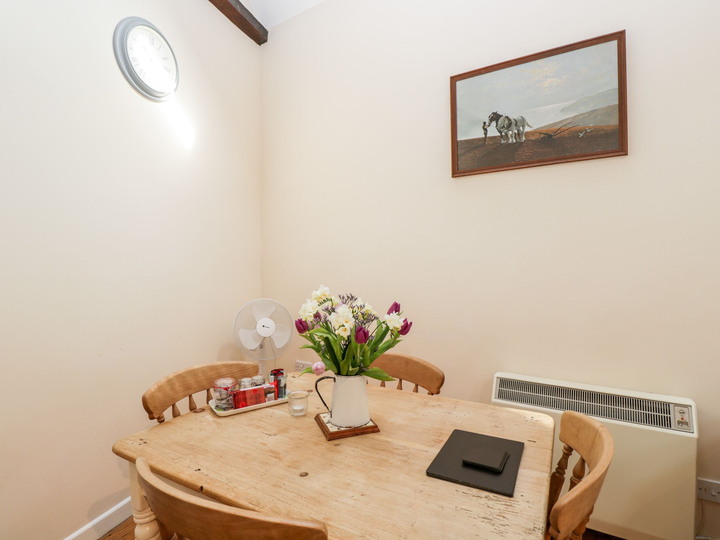 A dining area with a wooden table, chairs, a fan, a vase with flowers, a tray with jars, and a painting on the wall.