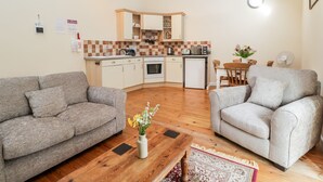 Un espacio habitable compacto con dos sofás grises, una mesa de centro de madera, una cocina con salpicadero de azulejos y una zona de comedor con mesa y sillas.