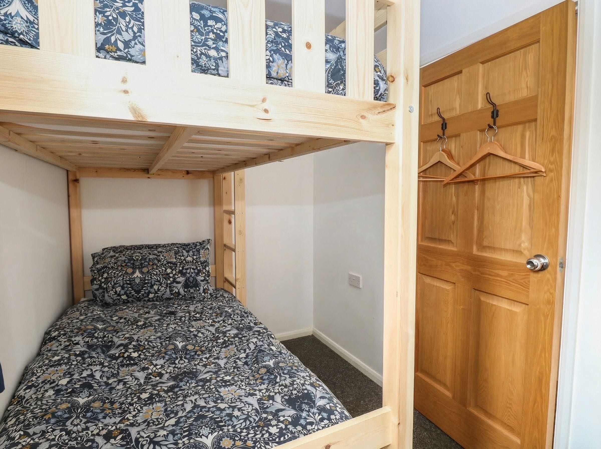 Ein Stockbett mit gemustertem Bettbezug, eine Holztür mit Kleiderhaken und ein Lichtschalter an der Wand.