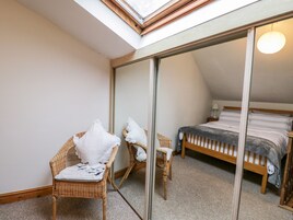 Un dormitorio con tragaluz, una cama con manta gris, dos sillas de mimbre con toallas blancas y un espejo grande que refleja la habitación.