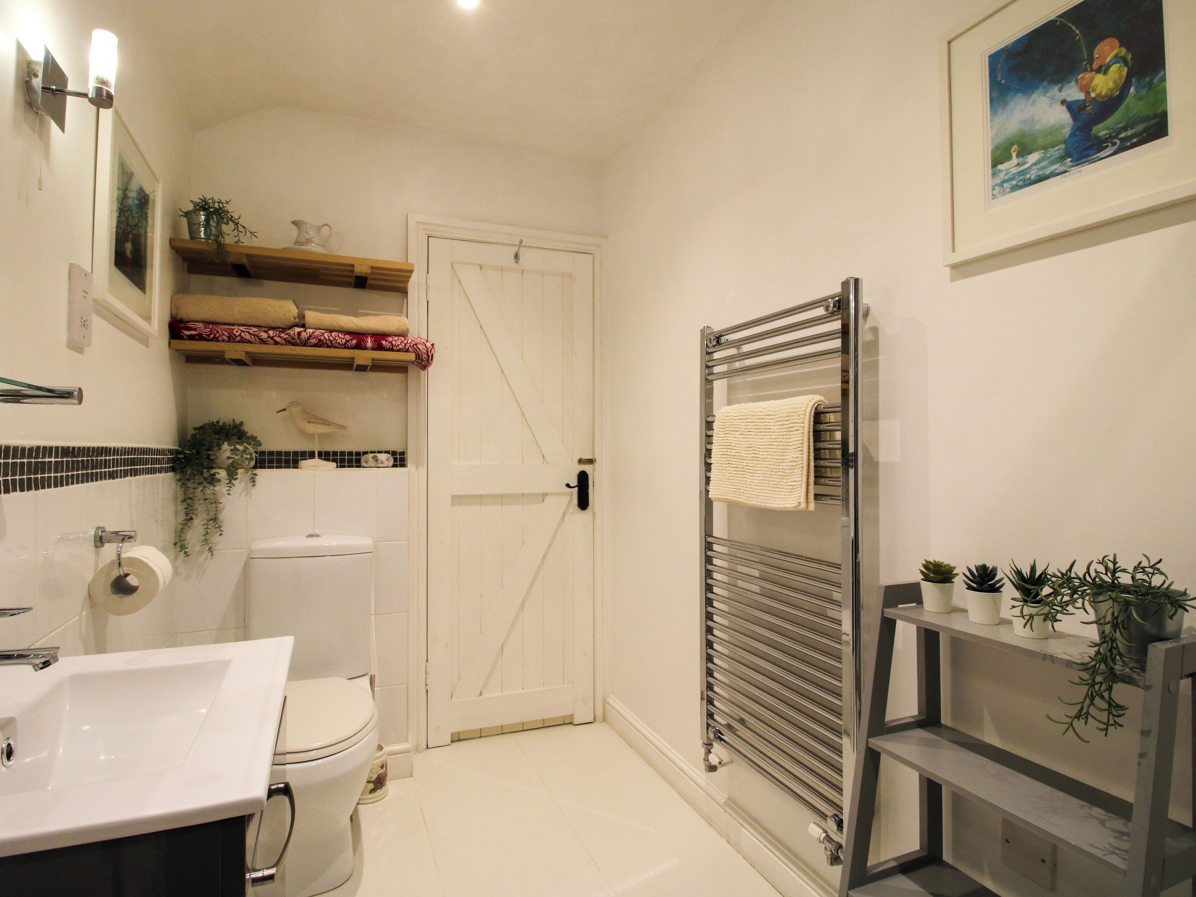 A bathroom with a white sink, toilet, and a glass shower enclosure. There are shelves with plants and a framed picture on the wall.