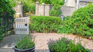 Un jardin avec un chemin de gravier, un portail en métal et deux plantes en pot.