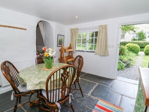 A dining area with a table set for four, surrounded by wooden chairs. A vase with flowers is on the table, and there is a window with curtains. - Hawthorn Cottage (Llandysul)