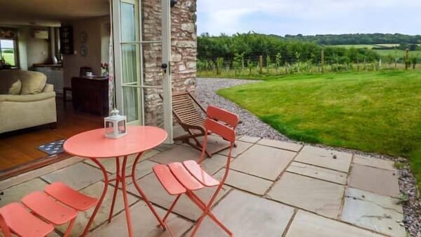 A patio area with a round table and chairs, a stone wall, and a view of a green field and hills. - Rose Cottage (Monmouth)