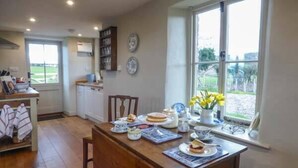 A dining area with a wooden table set for a meal, a window with a view of the outdoors, and a kitchen in the background. - Rose Cottage (Monmouth)