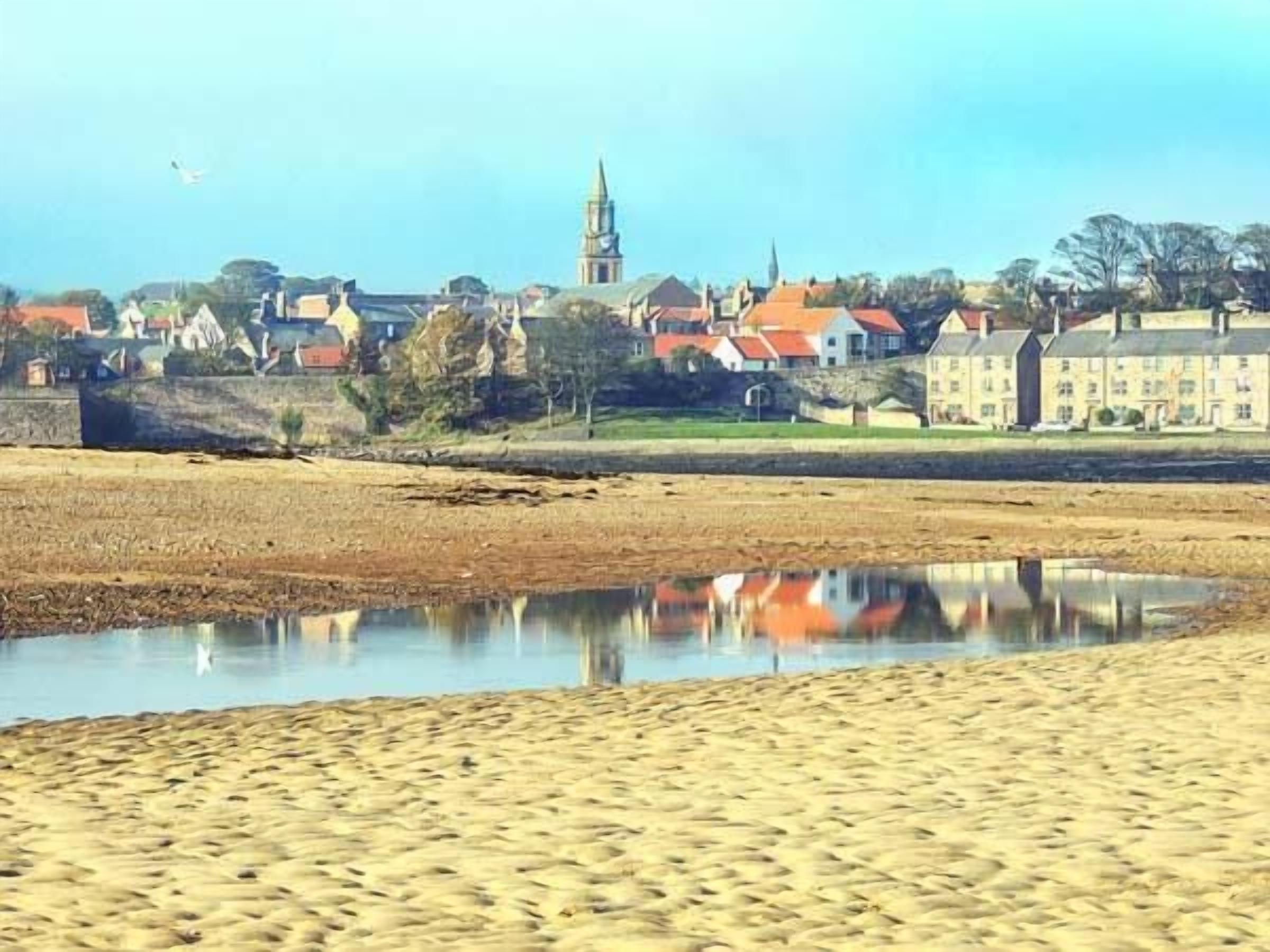Une plage de sable fin, avec une étendue d’eau calme qui reflète les bâtiments et le clocher d’une église en arrière-plan.