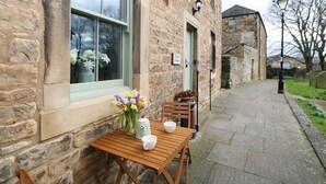 A stone building with a wooden table and chairs outside, a sign indicating 'Church Cottage', and a lamp post on the sidewalk.