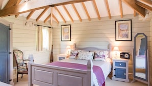 Une chambre avec un plafond en bois, un lit avec une tête de lit, des tables de chevet, une chaise, une commode et des œuvres d’art encadrées accrochées aux murs.