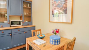 A compact kitchen with a microwave, a blue cabinet with glass doors, and a wooden dining table with a bowl of fruit and a vase of flowers. - The Garden Flat (Fort William)