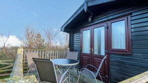 A wooden deck with a table and two chairs, a wooden fence, and a building with dark siding and frosted glass doors. - Sycamore Lodge (Saltburn-by-the-Sea)