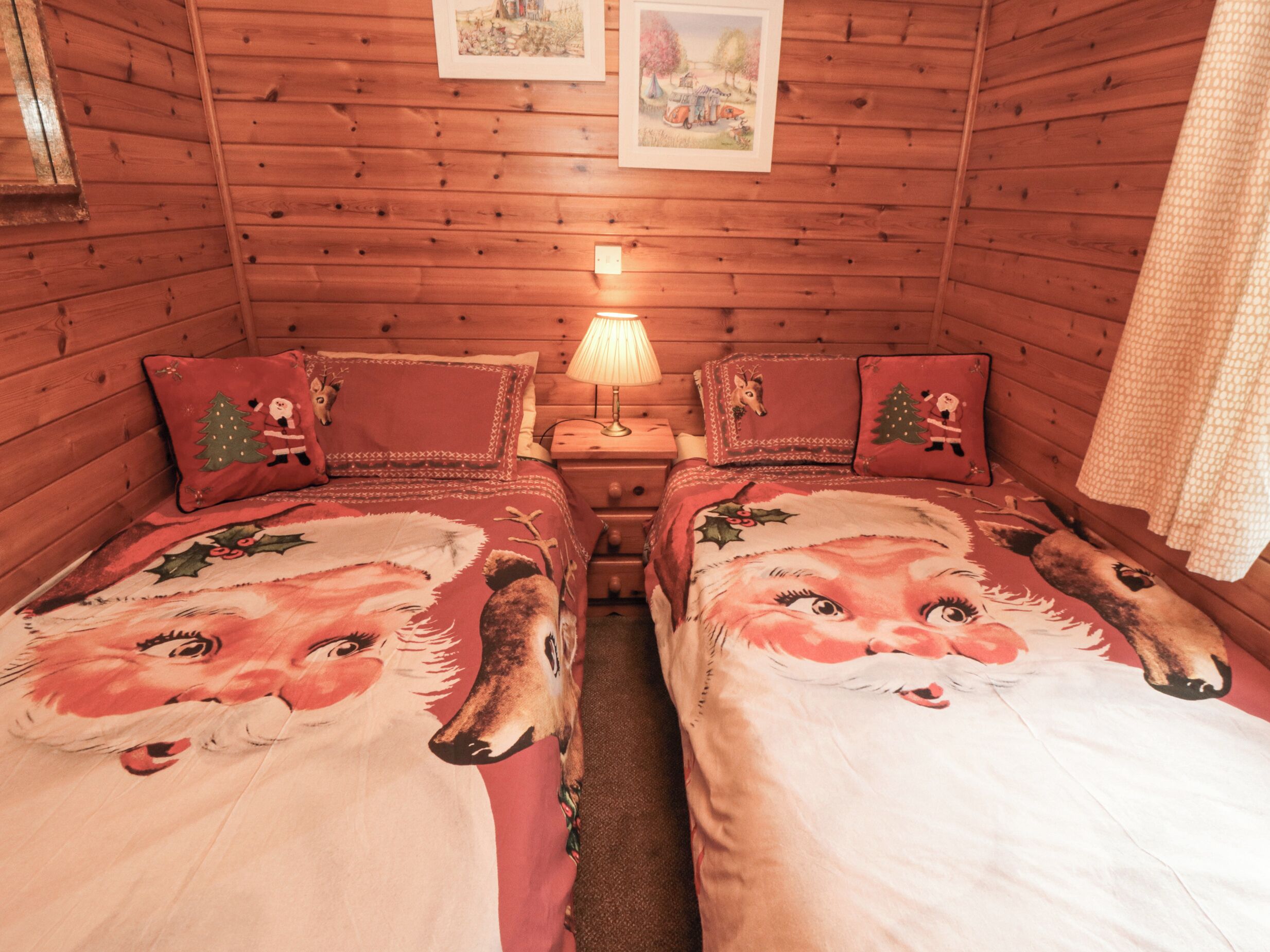 A cozy cabin room with two beds, each adorned with festive bedding featuring a reindeer and Christmas tree. A small table with a lamp and a picture frame is positioned between the beds.