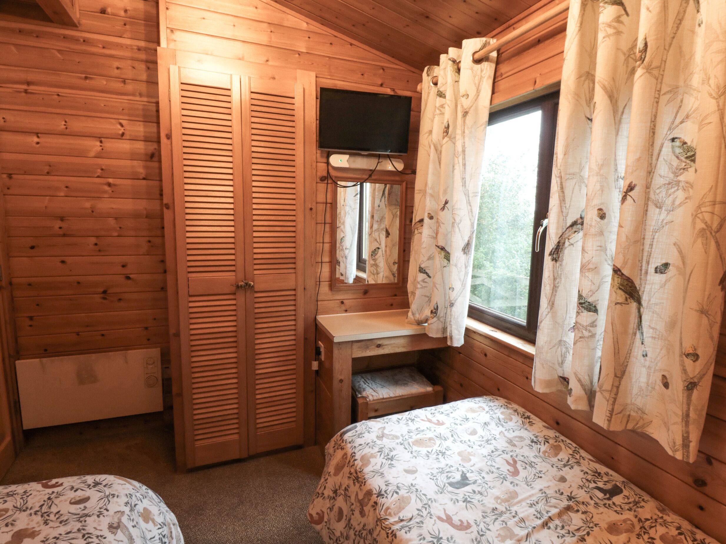 A wooden cabin room with two beds, a small desk, a television, and a window with curtains.