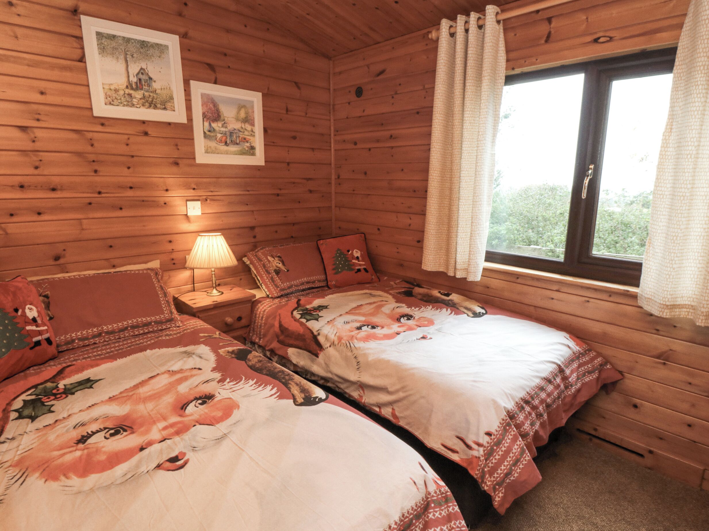A cozy room with wooden walls, two beds with festive bedding, a nightstand with a lamp, and a window with curtains.