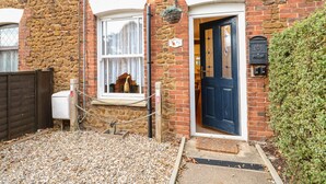 A stone building with a blue door, a window with lace curtains, and a small porch area. - Duck Cottage (King's Lynn)
