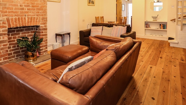 A cozy living room with a brick fireplace, leather sofas, and wooden flooring. - Duck Cottage (King's Lynn)
