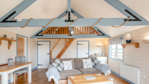 Ein gemütliches Zimmer mit Holzboden, einem weißen Sofa mit Karokissen, einem Holztisch und einer Treppe mit freiliegenden Balken und Seil.