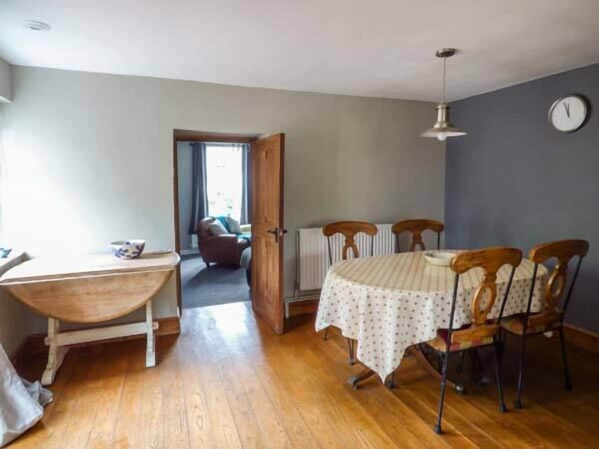 A dining area with a table and chairs, a clock on the wall, and a view into another room.