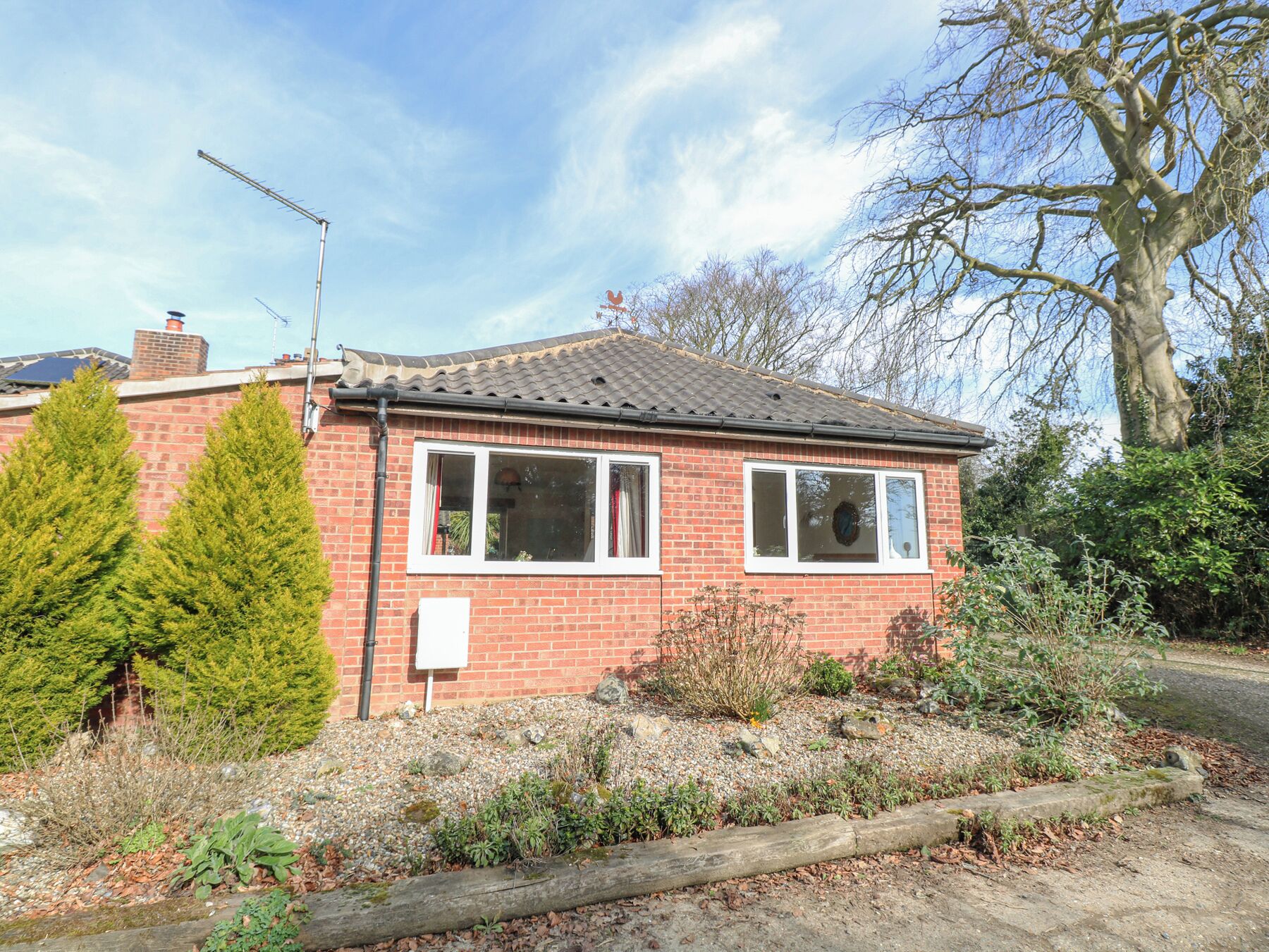A single-story brick house with a tiled roof, a chimney, and a small garden with various plants and a wooden fence.